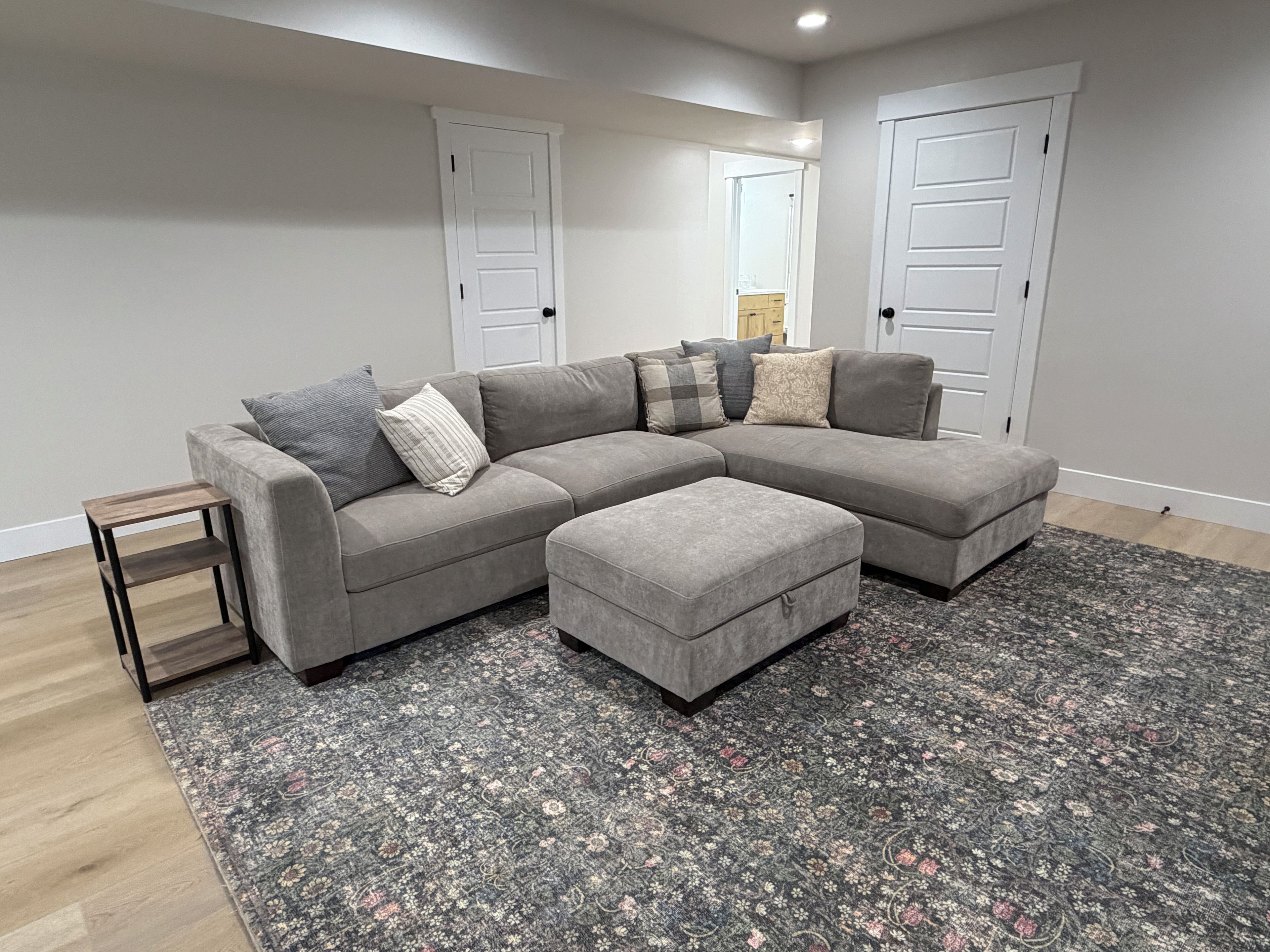 A grey sectional sofa with multiple cushions, a matching ottoman, a small wooden side table, and a patterned rug on a wooden floor.