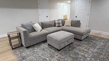 A grey sectional sofa with multiple cushions, a matching ottoman, a small wooden side table, and a patterned rug on a wooden floor.