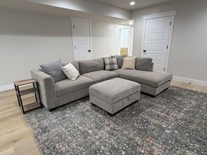 A grey sectional sofa with multiple cushions, a matching ottoman, a small wooden side table, and a patterned rug on a wooden floor. - Just-Finished 2BR Basement Suite • Private Entrance (Ammon)