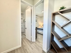 Un pasillo con suelo de madera, una puerta blanca y una cama con lámpara al fondo.