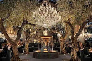 An outdoor dining area with a central fountain, surrounded by trees and illuminated by chandeliers.