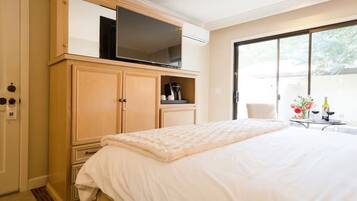 A bedroom with a large bed, a TV on a wooden cabinet, a small table with drinks, and a sliding glass door leading to a balcony.