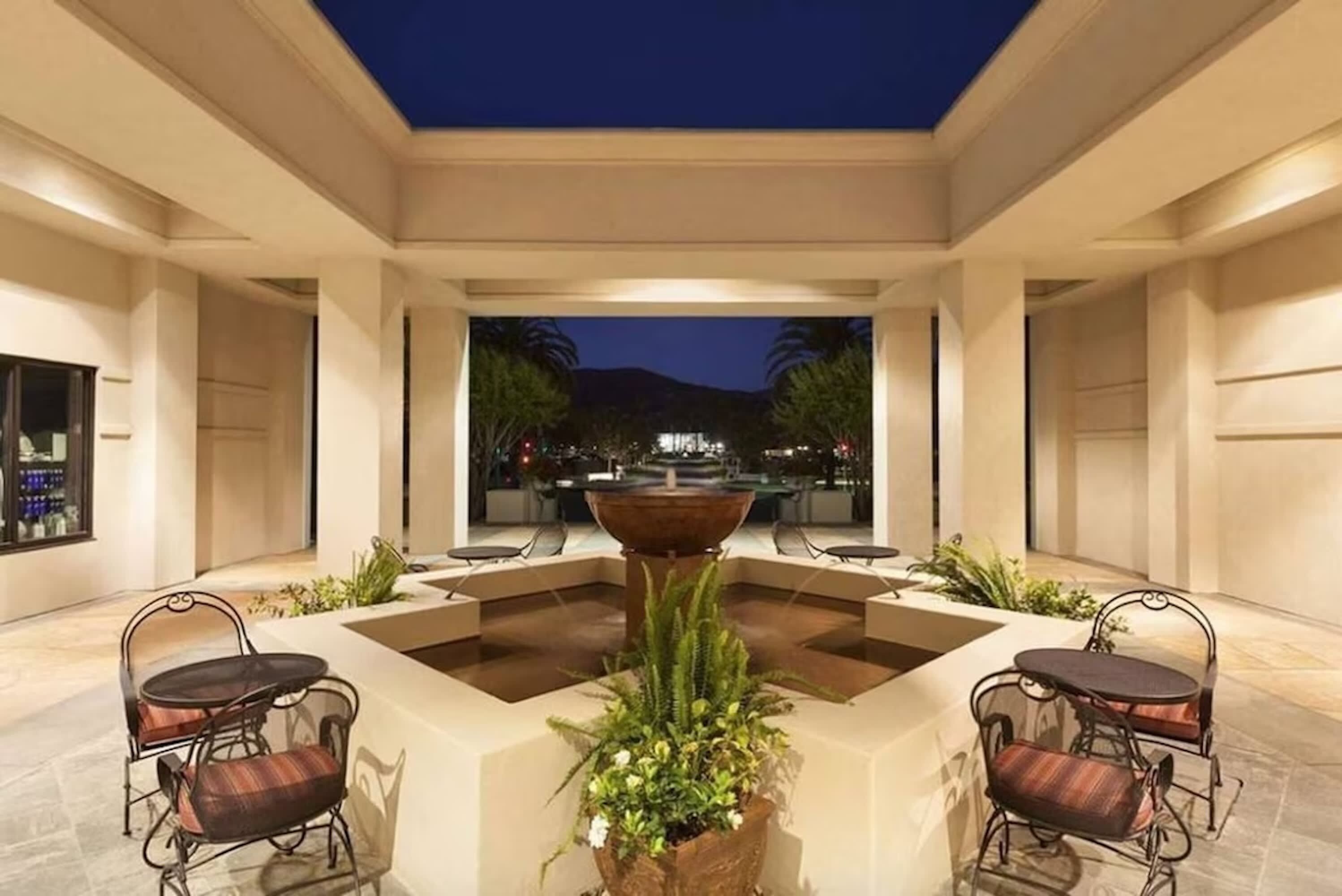 Un espace extérieur bien éclairé, aménagé en patio, avec une fontaine centrale, entouré de sièges et de plantes.