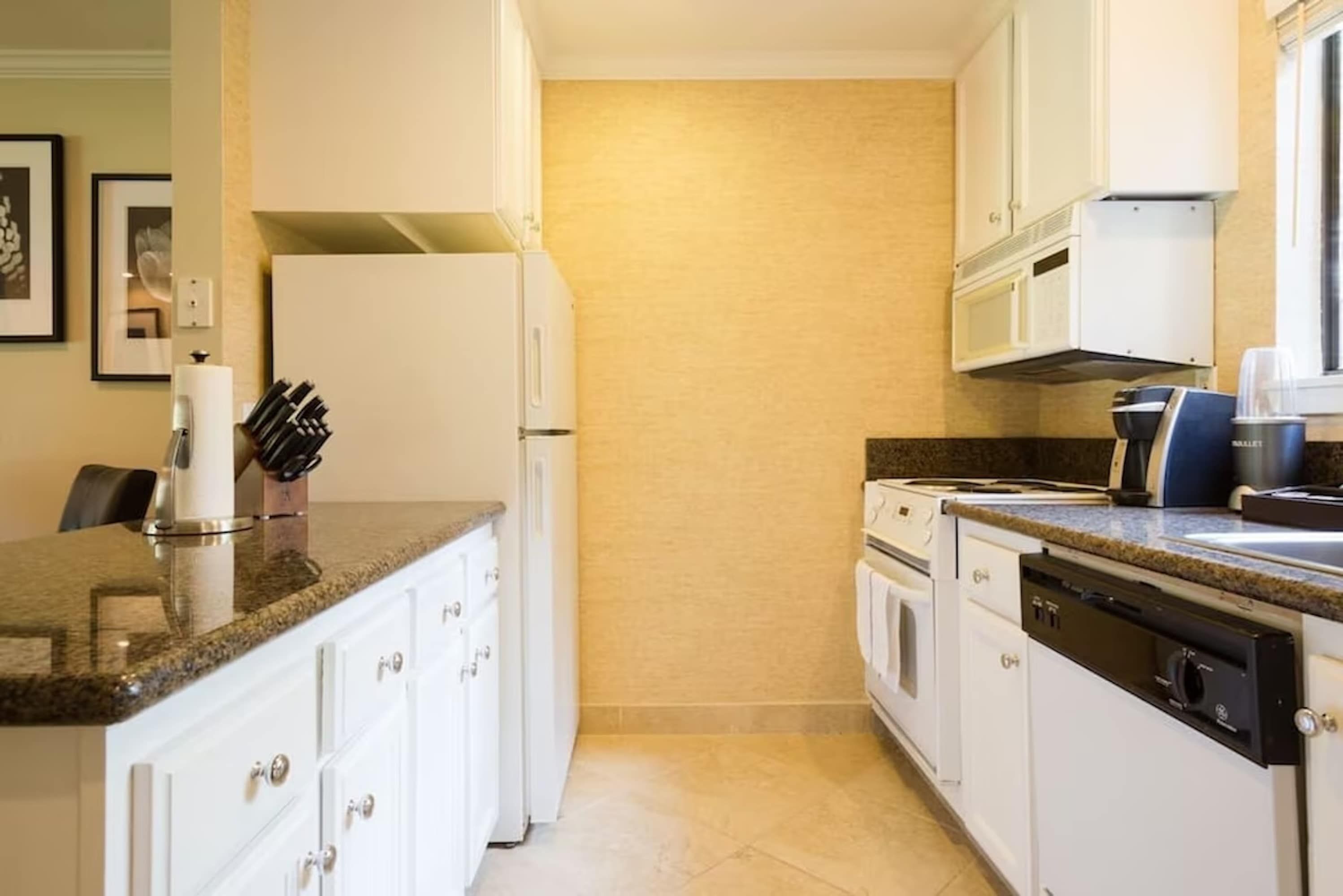 Una cocina con armarios blancos, encimera de granito, lavavajillas y cafetera.
