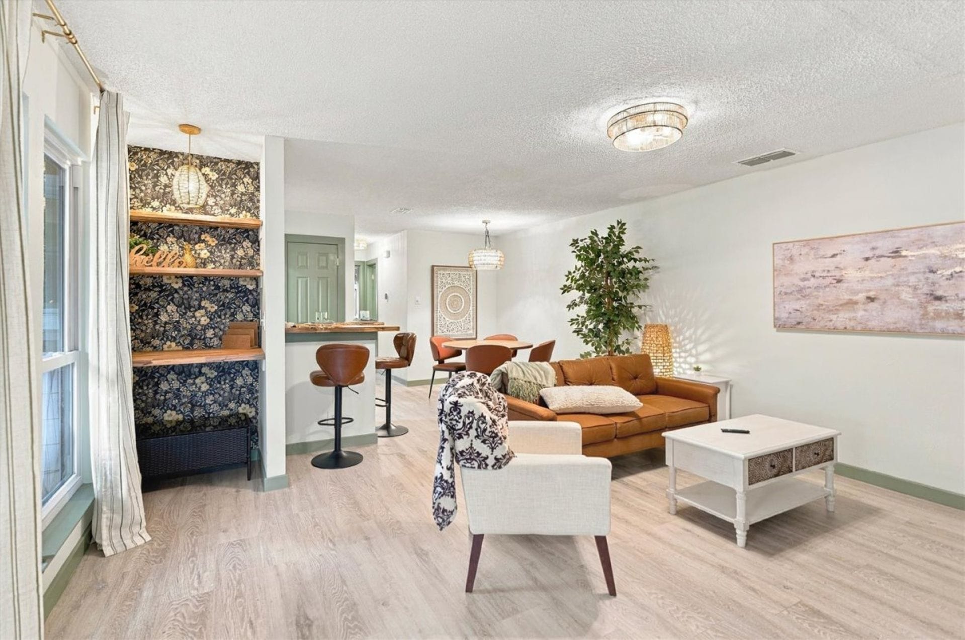 Settle into a warm, spacious living room with a caramel-toned sofa, comfortable seating, and soft natural light. Floral shelving, greenery, and layered artwork add character while the open layout connects naturally to the dining area.