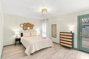 Unwind in a spacious, softly styled bedroom with a neatly made bed, gentle lighting, and calming neutral tones. Woven accents, framed décor, and the glass door add warmth and character to this peaceful retreat. - 2BR/2BA Home | Near The Domain & Apple (Austin)
