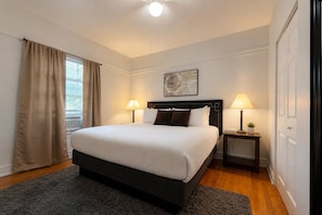 A hotel room with a large bed, two bedside lamps, a small table with a plant, a painting on the wall, and a window with curtains.