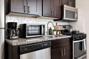 Una cocina con armarios oscuros, un microondas de acero inoxidable, una cafetera, una estufa y un lavavajillas.