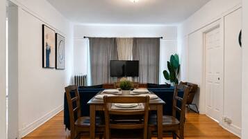 Uma sala de jantar com mesa posta para quatro pessoas, sofá, televisão e planta.
