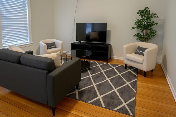 A living room with a sofa, two armchairs, a coffee table, a TV on a stand, a plant, and a window with blinds. - Fully Set Up 3BR w/ Big Kitchen (Chicago)