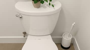 A white toilet with a plant on top, a toilet brush, and a bucket on the floor.
