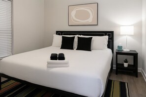 A neatly made bed with white and black bedding, a bedside lamp, a small potted plant, and a framed abstract artwork on the wall.