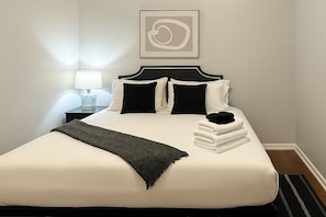 A neatly made bed with a black throw blanket, a bedside lamp, and a framed abstract artwork on the wall. - 3BR Apt - Prime & Spacious (Chicago)