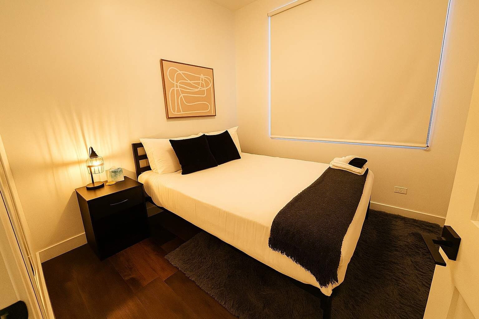 A neatly arranged bedroom with a bed, bedside table, lamp, and a wall-mounted artwork.