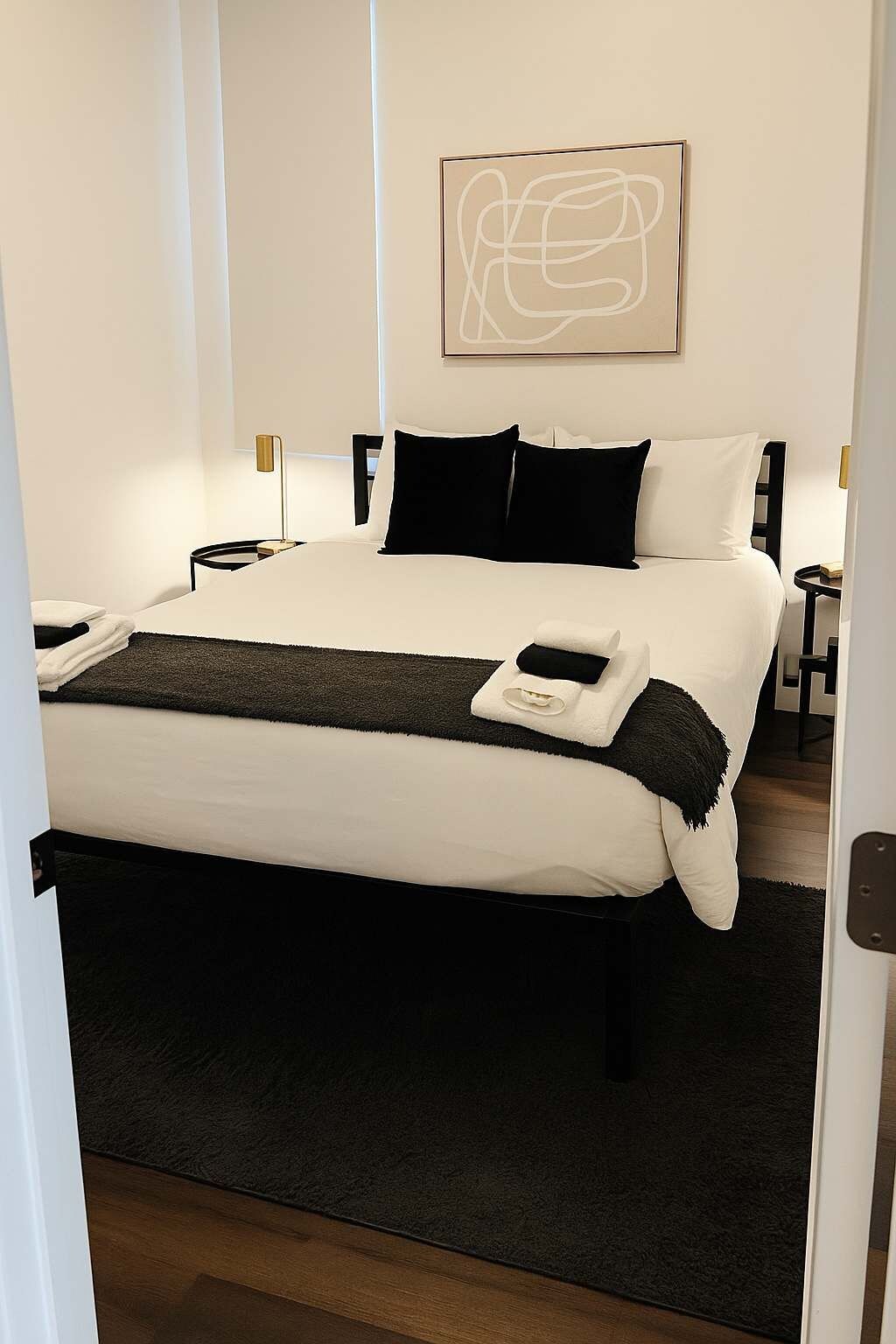 A modern hotel room with a large bed, black and white bedding, a black throw blanket, and a minimalist abstract painting on the wall.