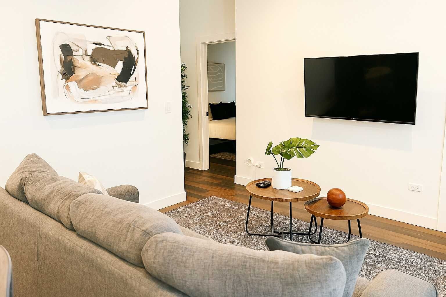A living room with a grey sofa, a round coffee table with a plant, a TV mounted on the wall, and a door leading to another room.