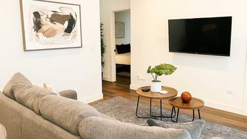 A living room with a grey sofa, a round coffee table with a plant, a TV mounted on the wall, and a door leading to another room.