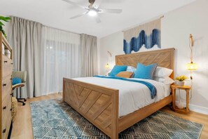 Un dormitorio con cama de madera, ventilador de techo, mesita de noche con lámpara y ventana con cortinas.