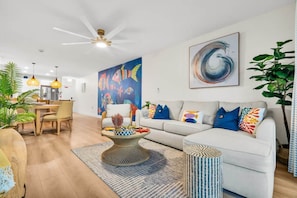 Ein modernes Wohnzimmer mit einem weißen Sofa, einem Esstisch mit Stühlen, einem Deckenventilator und einem großen abstrakten Gemälde.