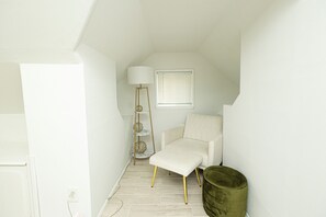 A small, white-walled room with a white chair, a floor lamp, and a window with blinds.