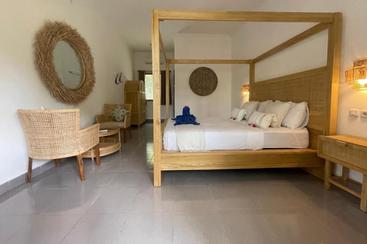 Quarto com cama de dossel, cadeira de vime, espelho redondo e luminária fixada na parede.