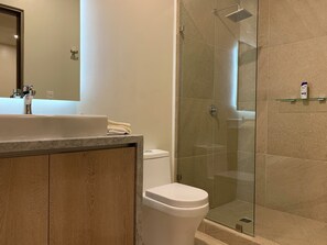 Casa de banho com chuveiro de vidro, sanita branca e bancada de mármore com lavatório.