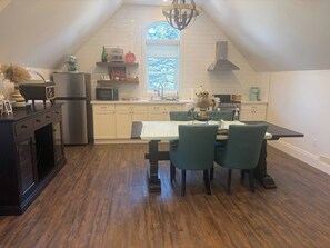 A modern kitchen with a dining area, featuring a wooden floor, white cabinetry, and a large window providing natural light.