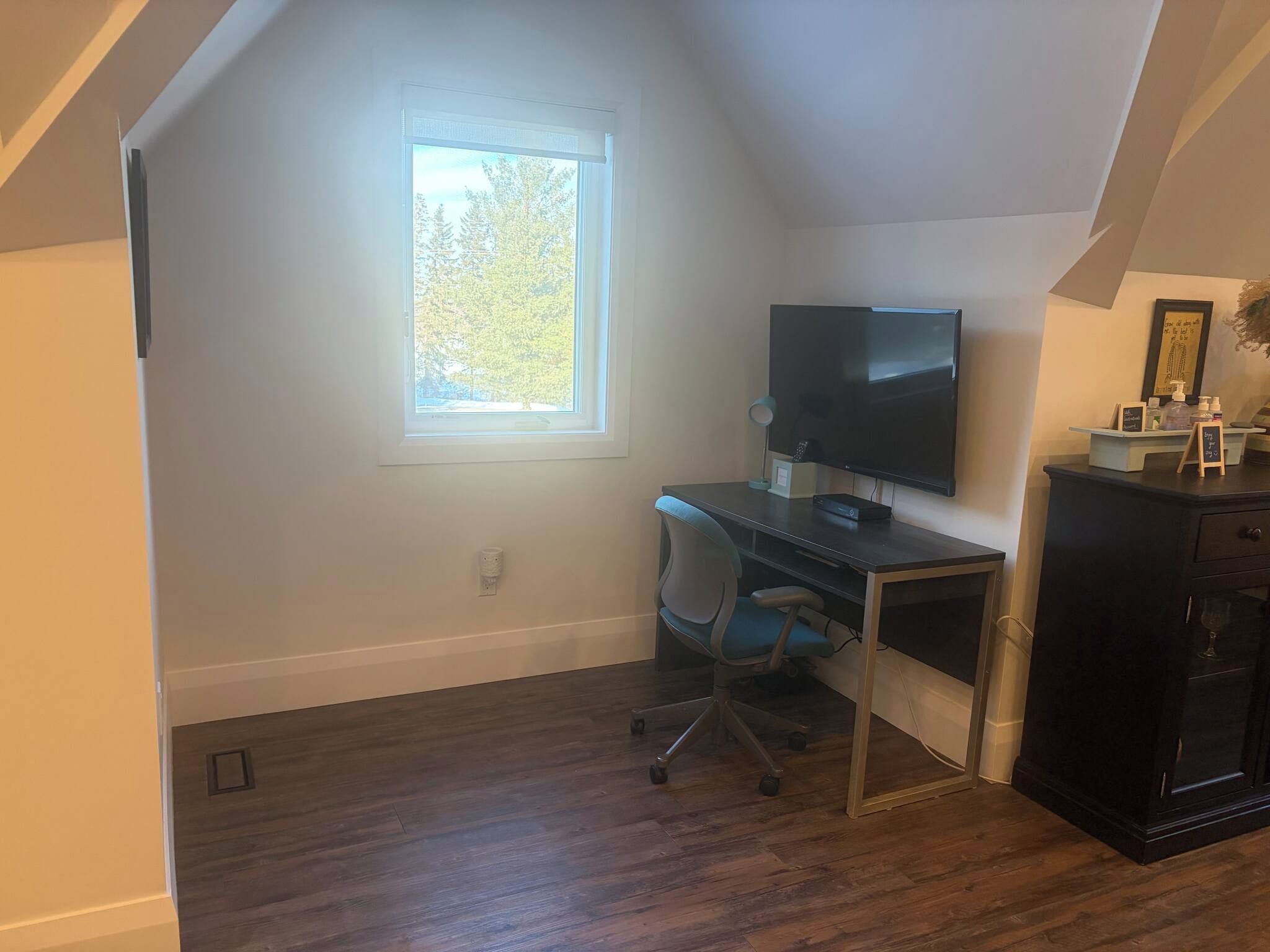 Une pièce comprenant un bureau, une chaise, un téléviseur et une fenêtre donnant sur des arbres.