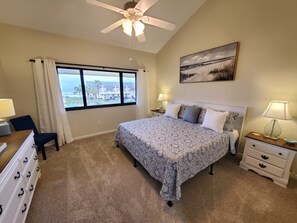 A bedroom with a bed, a desk, a chair, a nightstand, a lamp, a ceiling fan, a window with curtains, and a painting on the wall. - Summerhouse 147 2 Bedroom Condo (St. Augustine)