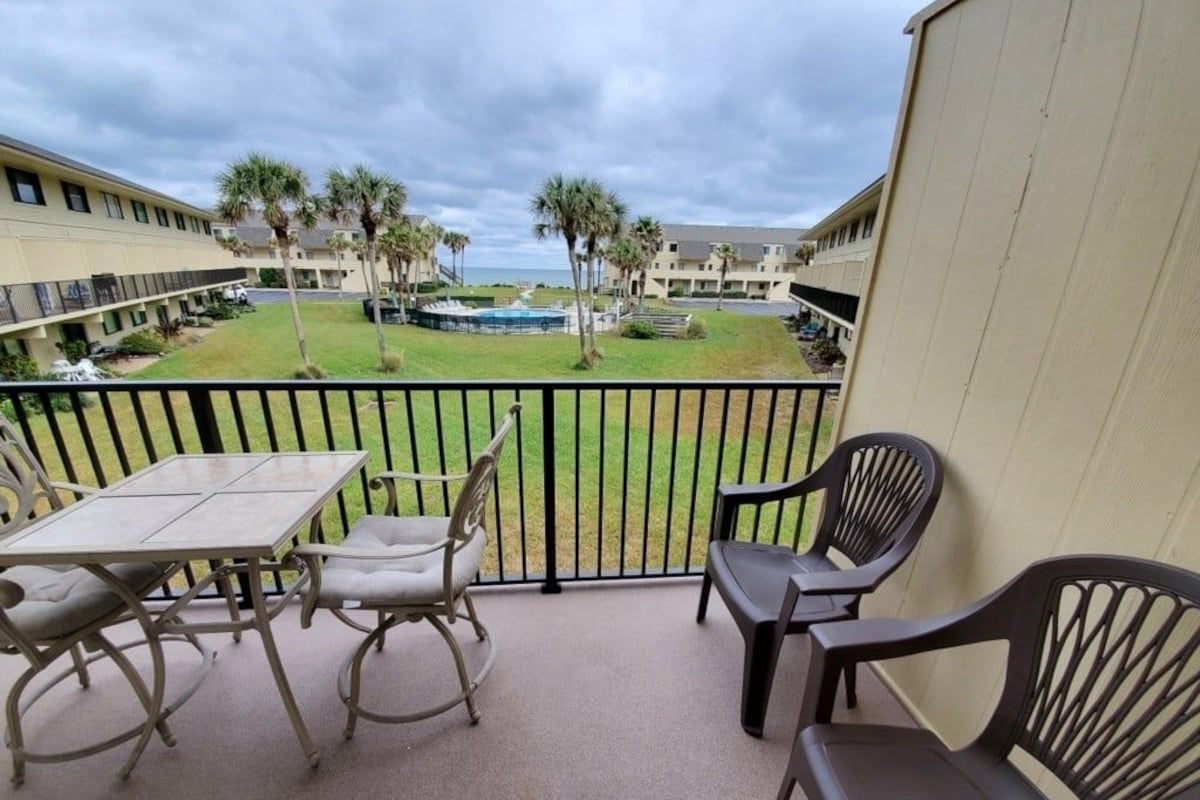 A balcony with a table and chairs, overlooking a pool and a lawn with palm trees.