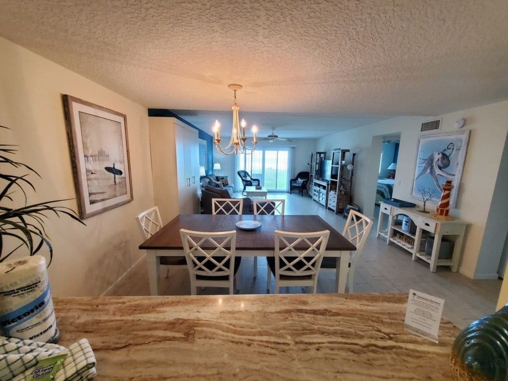 A dining area with a marble table and white chairs, a chandelier, and a view into the living room.