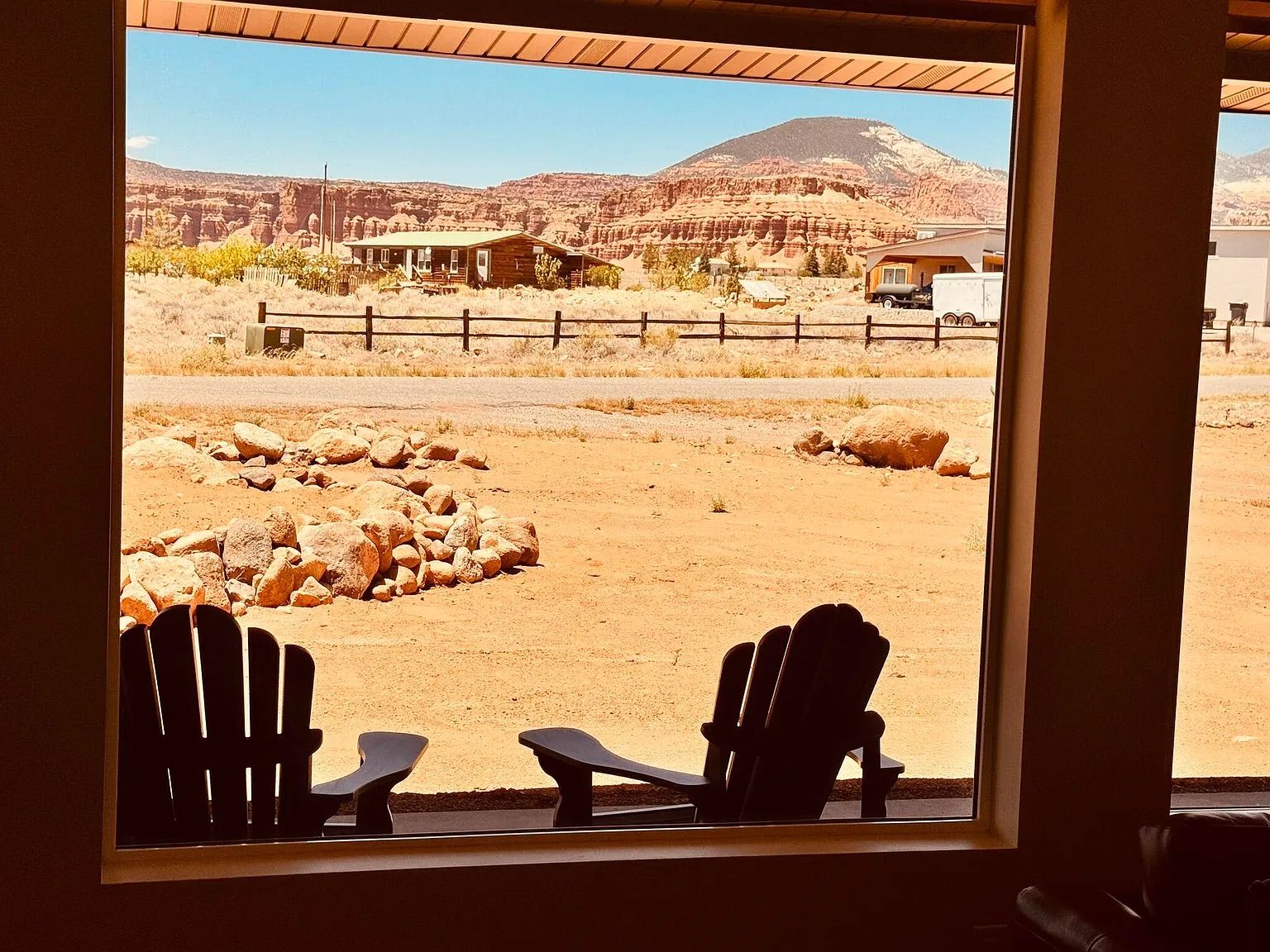 Blick aus dem Fenster auf eine Wüstenlandschaft mit einem Berg, einem Holzzaun und zwei Adirondack-Stühlen.