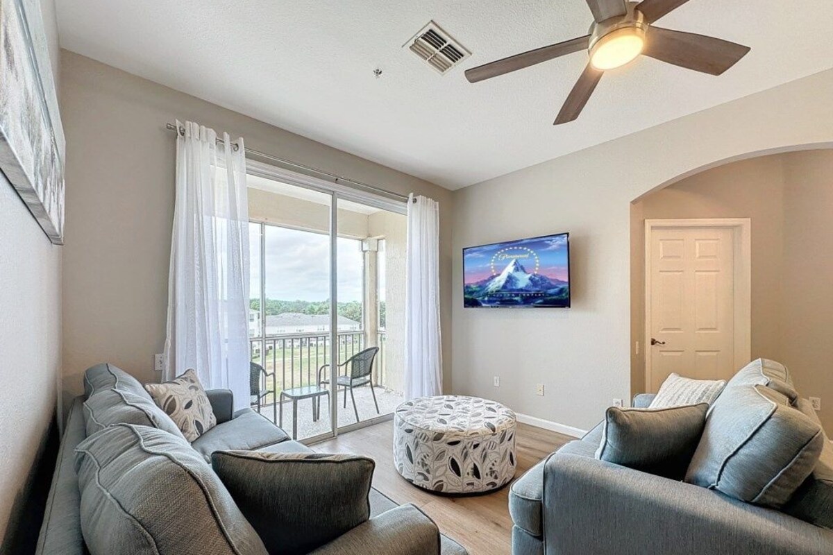 A living room with a sofa set, a round ottoman, a ceiling fan, and a television mounted on the wall.