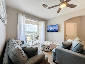 A living room with a sofa set, a round ottoman, a ceiling fan, and a television mounted on the wall. - Windsor Palms 3 Bed Condo In Gated Resort 3 Bedroom Condo (Kissimmee)