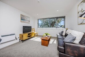 A modern living room with a flat-screen TV, a wooden entertainment unit, a green plant, a leather sofa, and a large window with a view of trees. - Eden Vista Apartment 3 Bedrooms with free parking (Auckland)