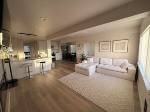 Una sala de estar moderna con un amplio sofá modular, una cocina con refrigerador y un comedor con sillas.