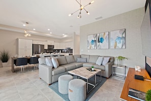 A modern living room with a grey sofa, ottomans, a coffee table, and a dining area with a wooden table and chairs. - Fireworks @ Night -storey Lake By Shine Villas #709 2 Bedroom Condo (Kissimmee)