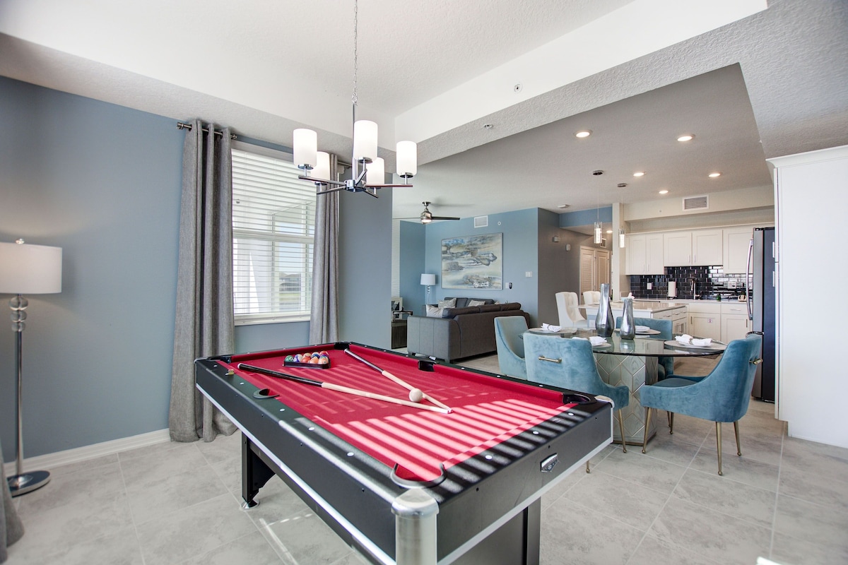 A pool table in a modern living area with a dining table and chairs, a kitchen in the background, and a ceiling fan.