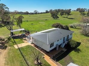 A single-story house with a metal roof, surrounded by a fenced yard and a green field.