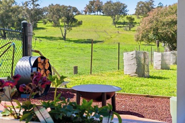 A barbecue grill with a metal chimney, a table, and a garden with a fence and trees in the background.