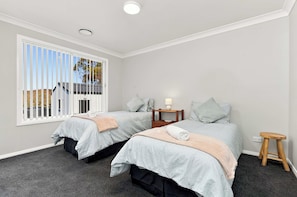 A bedroom with two single beds, a nightstand with a lamp, a small wooden stool, and a window with blinds.