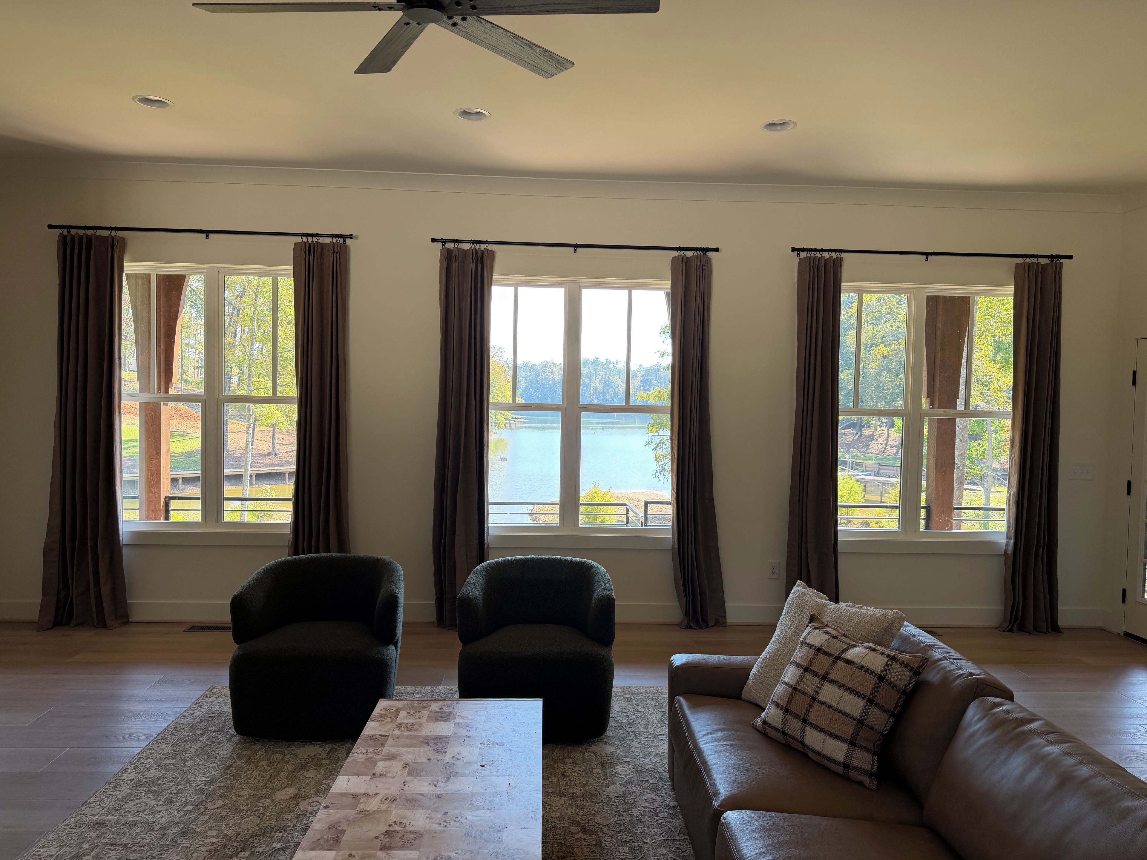 Ein geräumiges Wohnzimmer mit großem Fenster mit Seeblick, zwei Sessel, eine Couch und einem Deckenventilator.