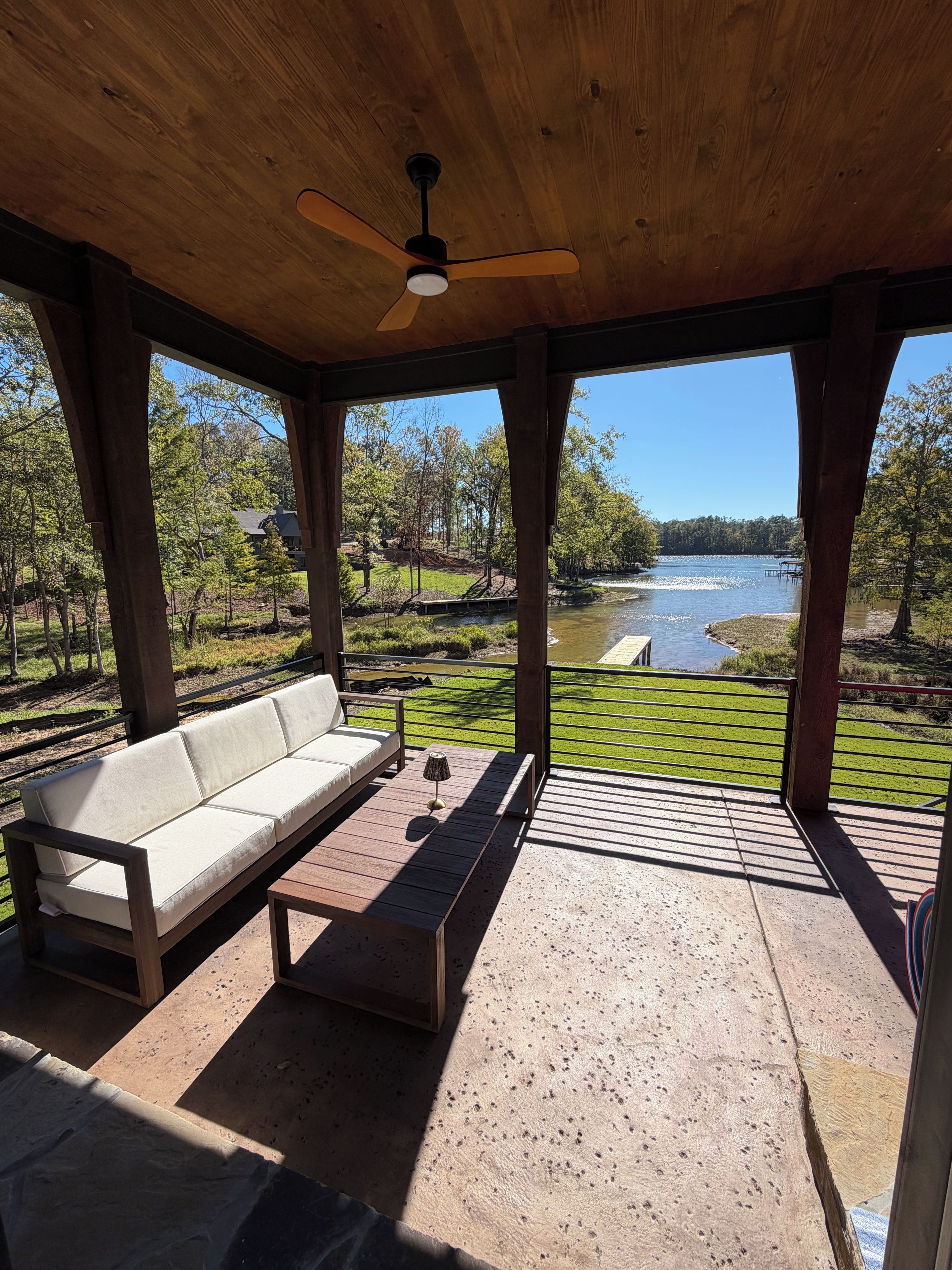 A covered outdoor seating area with a sofa and table, overlooking a lake and greenery.