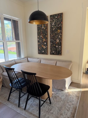 A dining area with a wooden oval table, black chairs, and a black pendant light. There are two floral paintings on the wall.