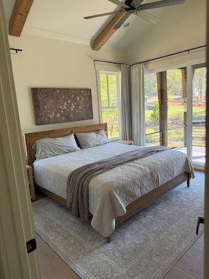 A bedroom with a large bed, a ceiling fan, a view of the outdoors, and a painting on the wall.