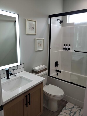 Ein modernes Badezimmer mit Waschbecken, Toilette und Dusche in Weiß. An den Wänden befinden sich Bilder in Rahmen und Duschutensilien auf der Duschablage.