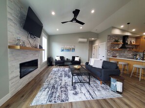 A modern living room with a fireplace, a flat-screen TV, a ceiling fan, and a kitchen area with bar stools. - Modern Coastal 3-bedroom Beach Retreat, Ocean Shores with full kitchen, Fire Pit (Ocean Shores)
