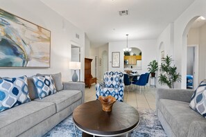 Een moderne woonkamer met een grijze bank, een ronde salontafel en gekleurde kussens. Er is een eethoek met blauwe stoelen en een keuken op de achtergrond.