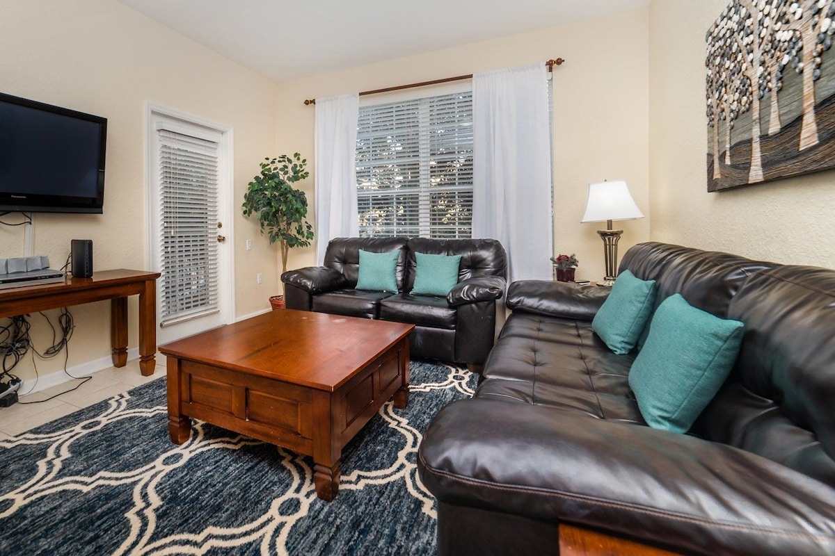 A living room with a flat-screen TV, leather sofas, a coffee table, a plant, and a window with blinds.