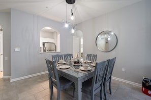 A dining area with a rectangular table set for six, surrounded by six chairs. A kitchen with a microwave and sink is visible in the background. - Cw7650-204 3 Bedroom Condo (Kissimmee)